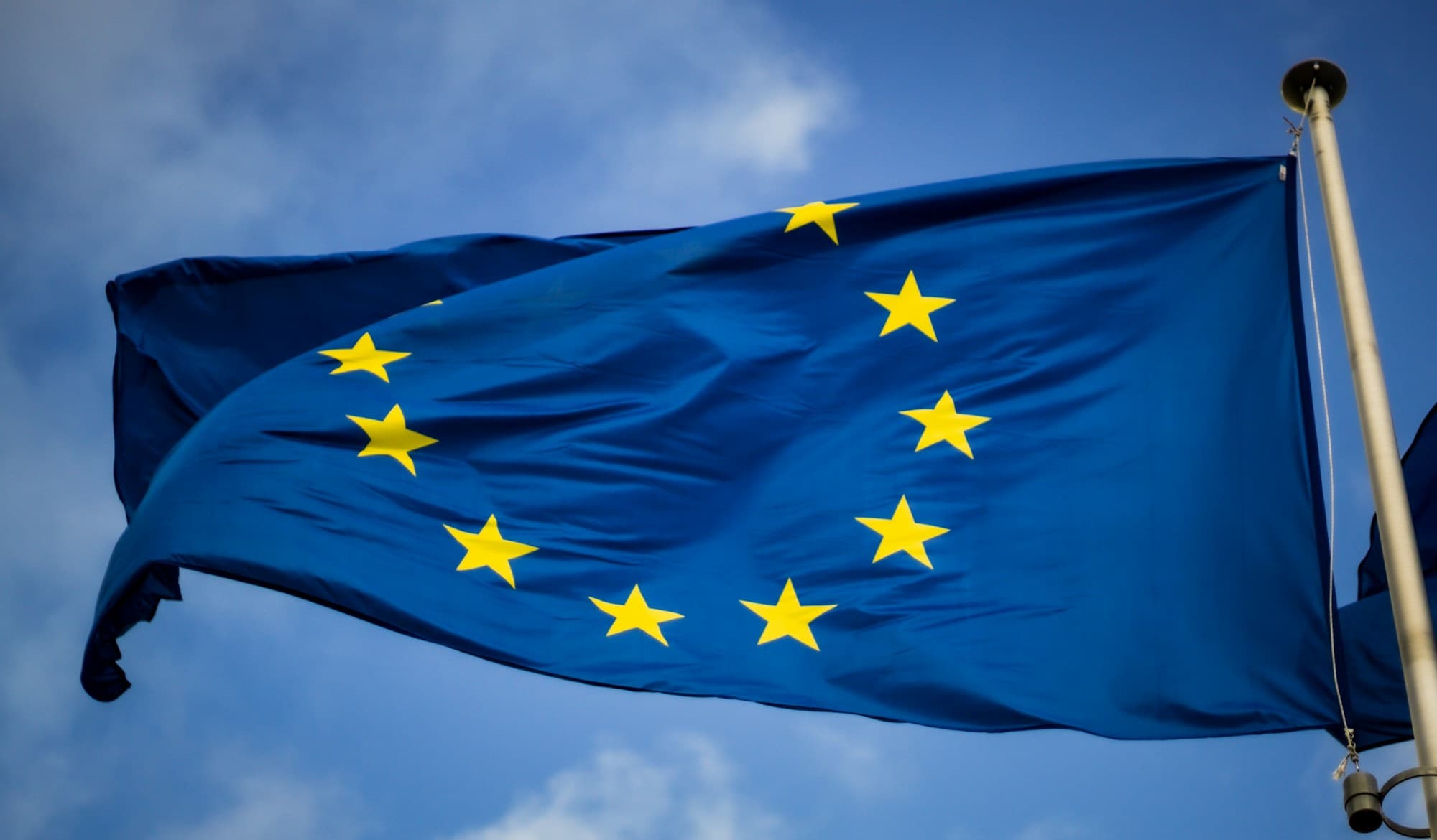 A European Union flag, a circle of gold stars on deep blue, against a clear sky.
