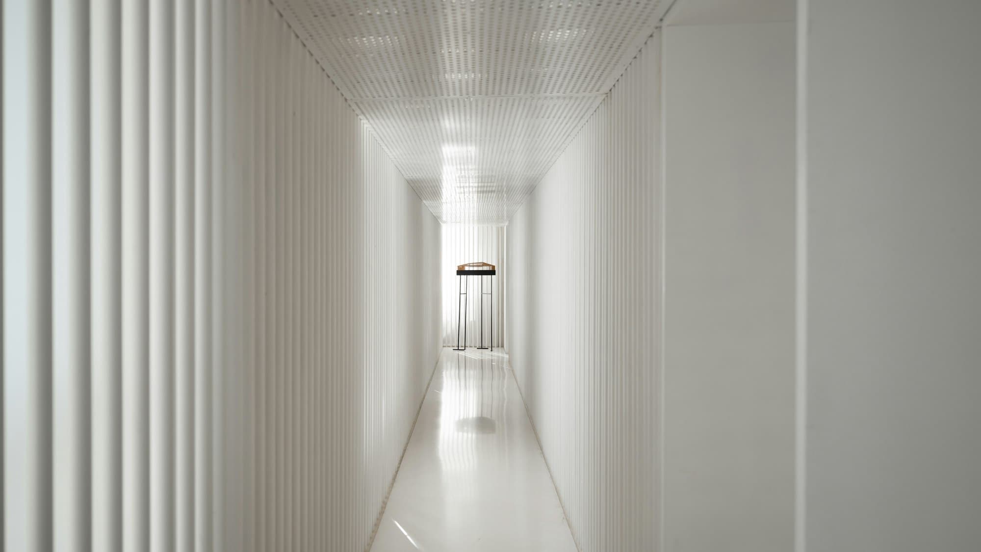 A clean white corridor in sharp one-point perspective, leading forward toward daylight.